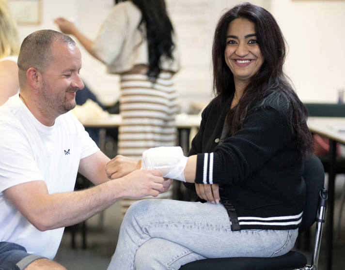 A man kneeling and wrapping a bandage around a woman's forearm during a first aid training session in a classroom setting, with other participants in the background. A man kneeling and wrapping a bandage around a woman's forearm during a first aid training session in a classroom setting, with other participants in the background.